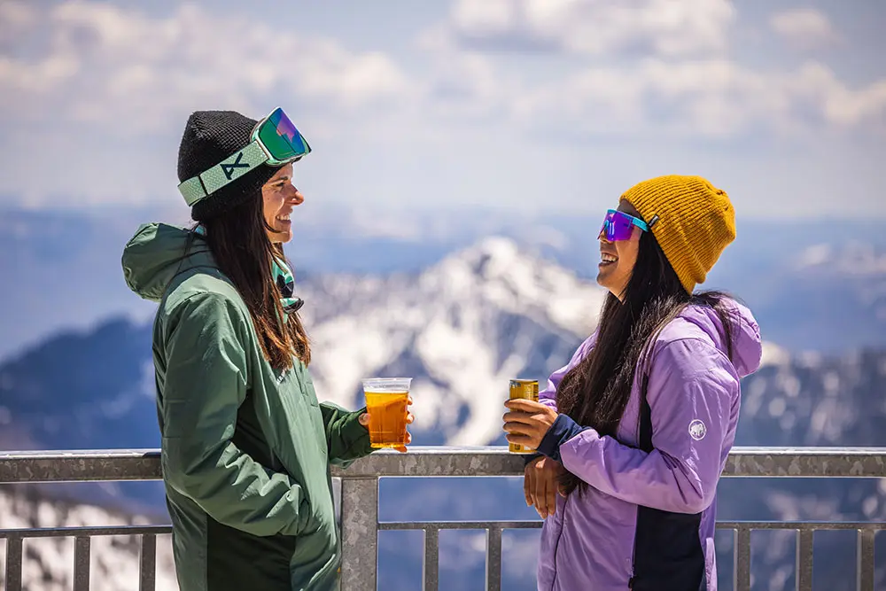 2 friends laugh while holding beers next to the balcony at The Summit, snowbird's mountain-top lunch restaurant